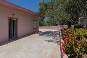 A building with a flat roof, a door, and a window. There is a tree and a flower bed with pink flowers. - Palm Beach House (Kissamos)
