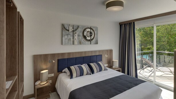 A modern bedroom with a large bed, wooden headboard, bedside tables with lamps, and a view of the outdoors through large windows. - Hotel Restaurant Le Domaine des Oliviers (Pierrelatte)