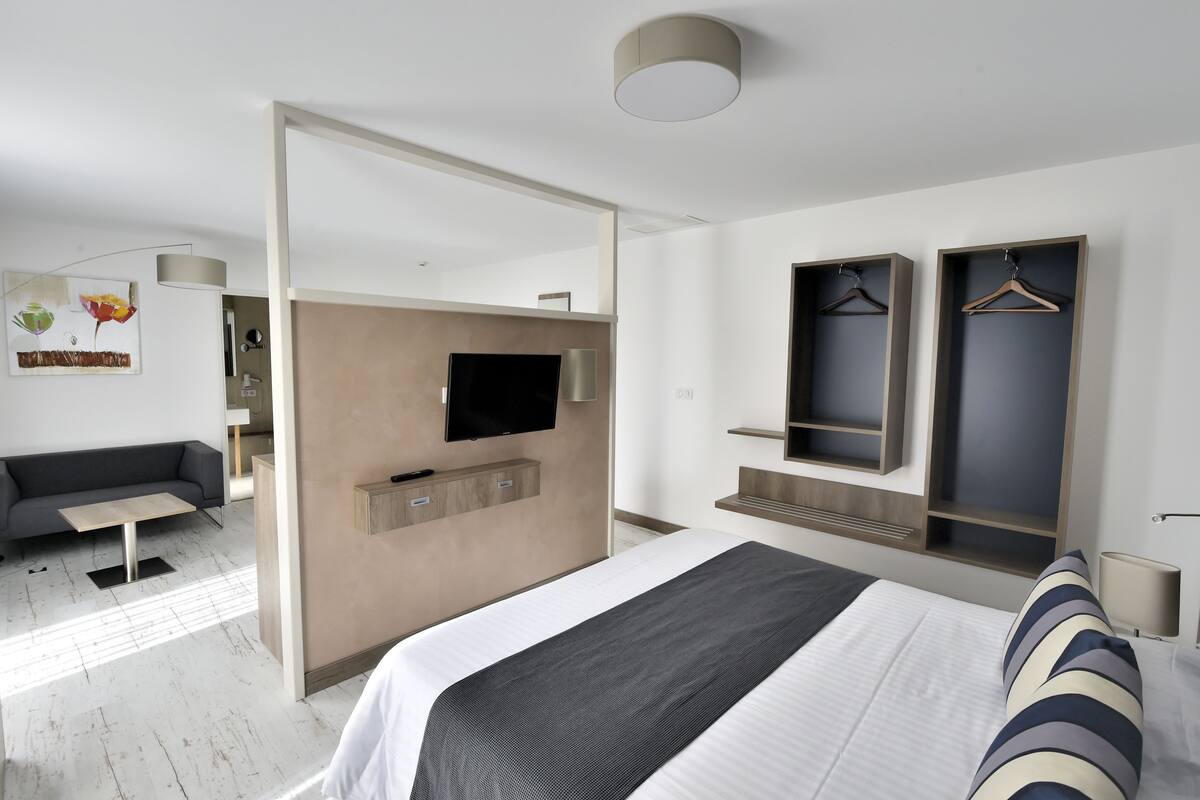 A modern hotel room with a large bed, a sofa, a TV mounted on the wall, and built-in wardrobes.