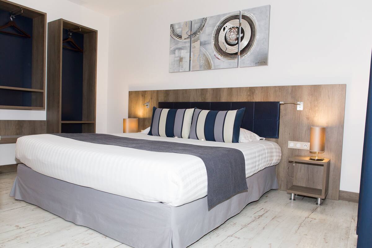 A modern hotel room with a large bed, a wooden headboard, two bedside tables with lamps, and a wall-mounted abstract art piece.