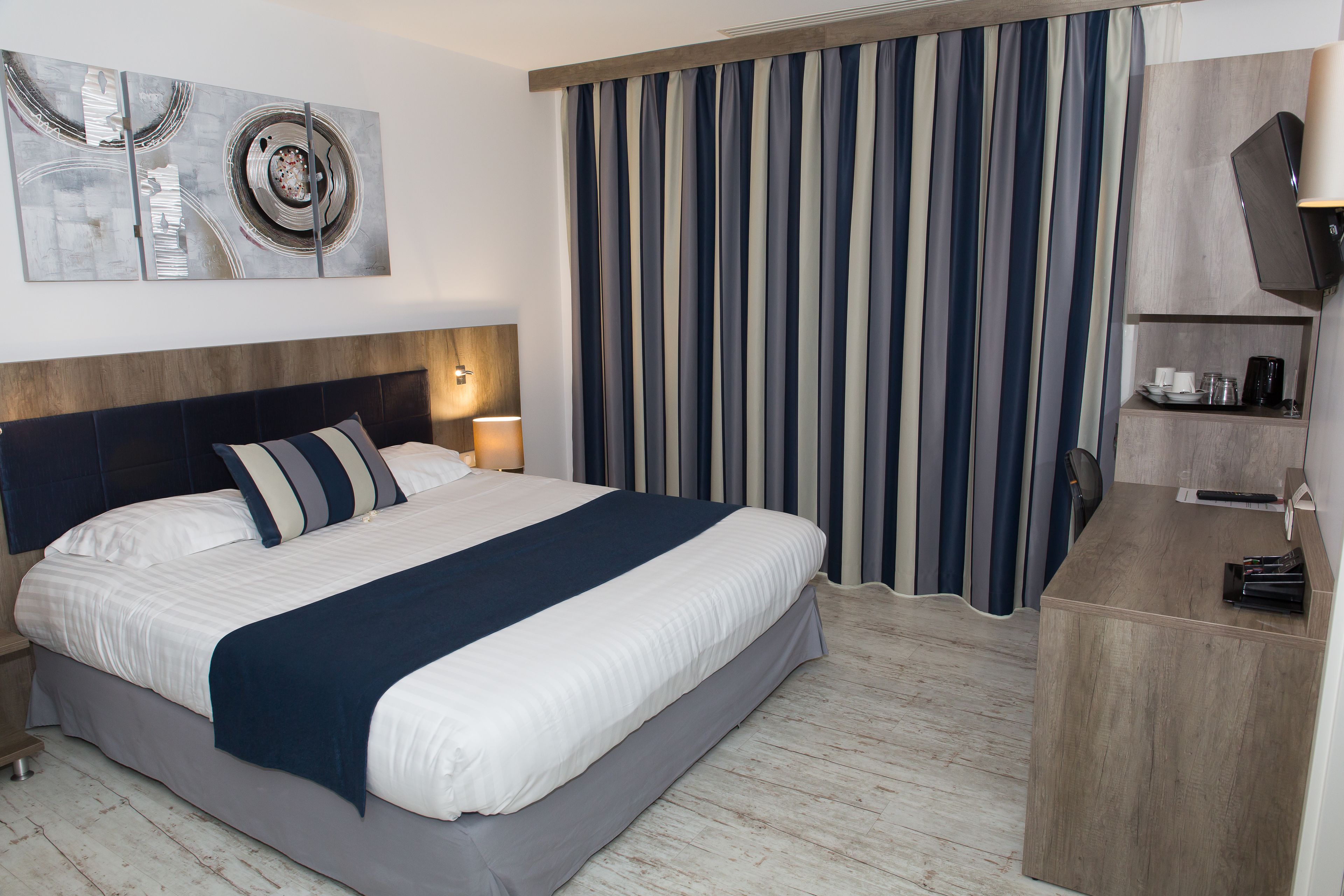 Une chambre d’hôtel avec un grand lit, une tête de lit, une table de chevet avec une lampe, un bureau avec une chaise, une télévision et un rideau à motifs rayés.