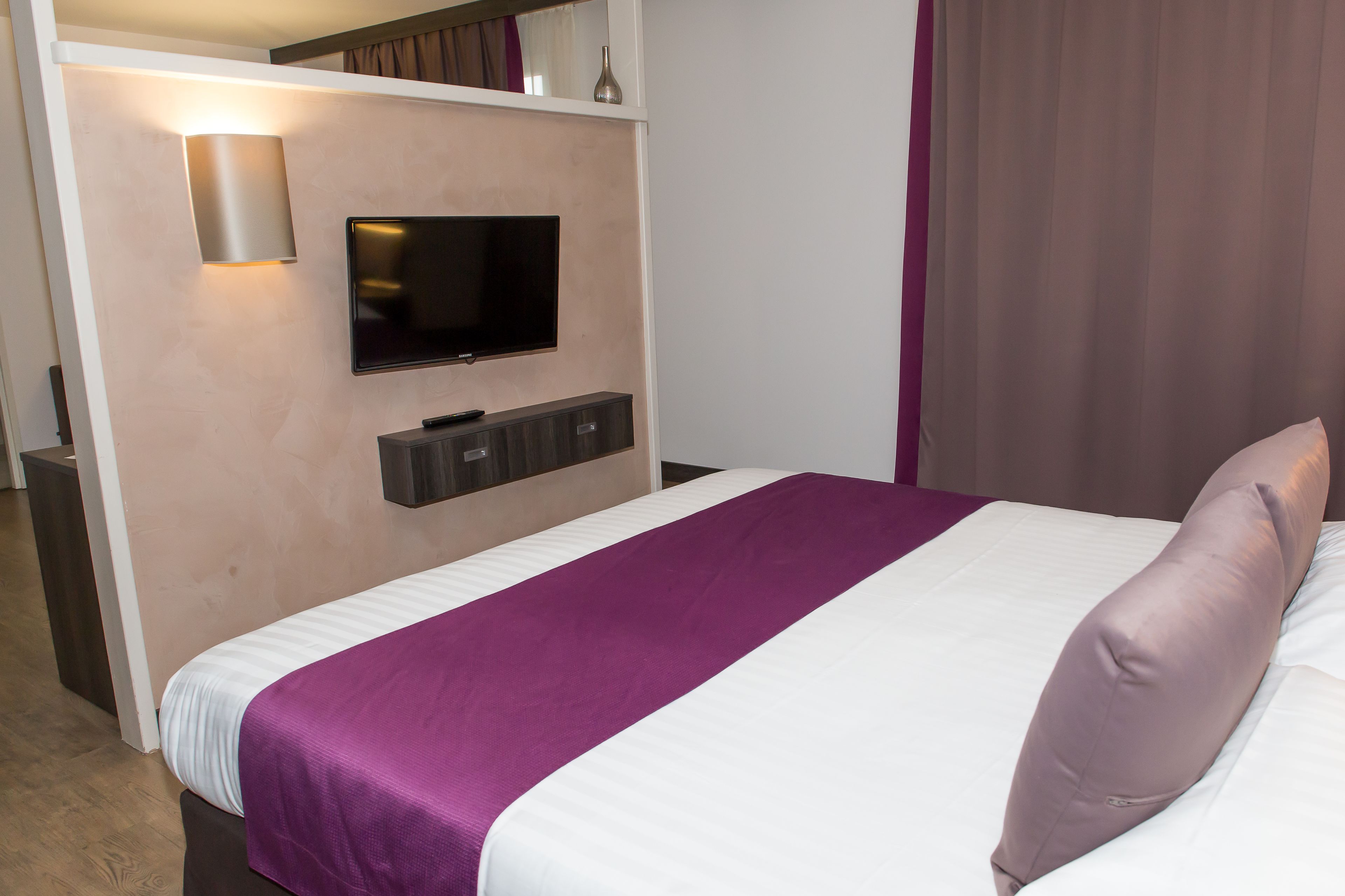 Habitación de hotel con cama, televisión instalada en la pared y una pequeña estantería sobre la cama.