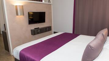 Habitación de hotel con cama, televisión instalada en la pared y una pequeña estantería sobre la cama.
