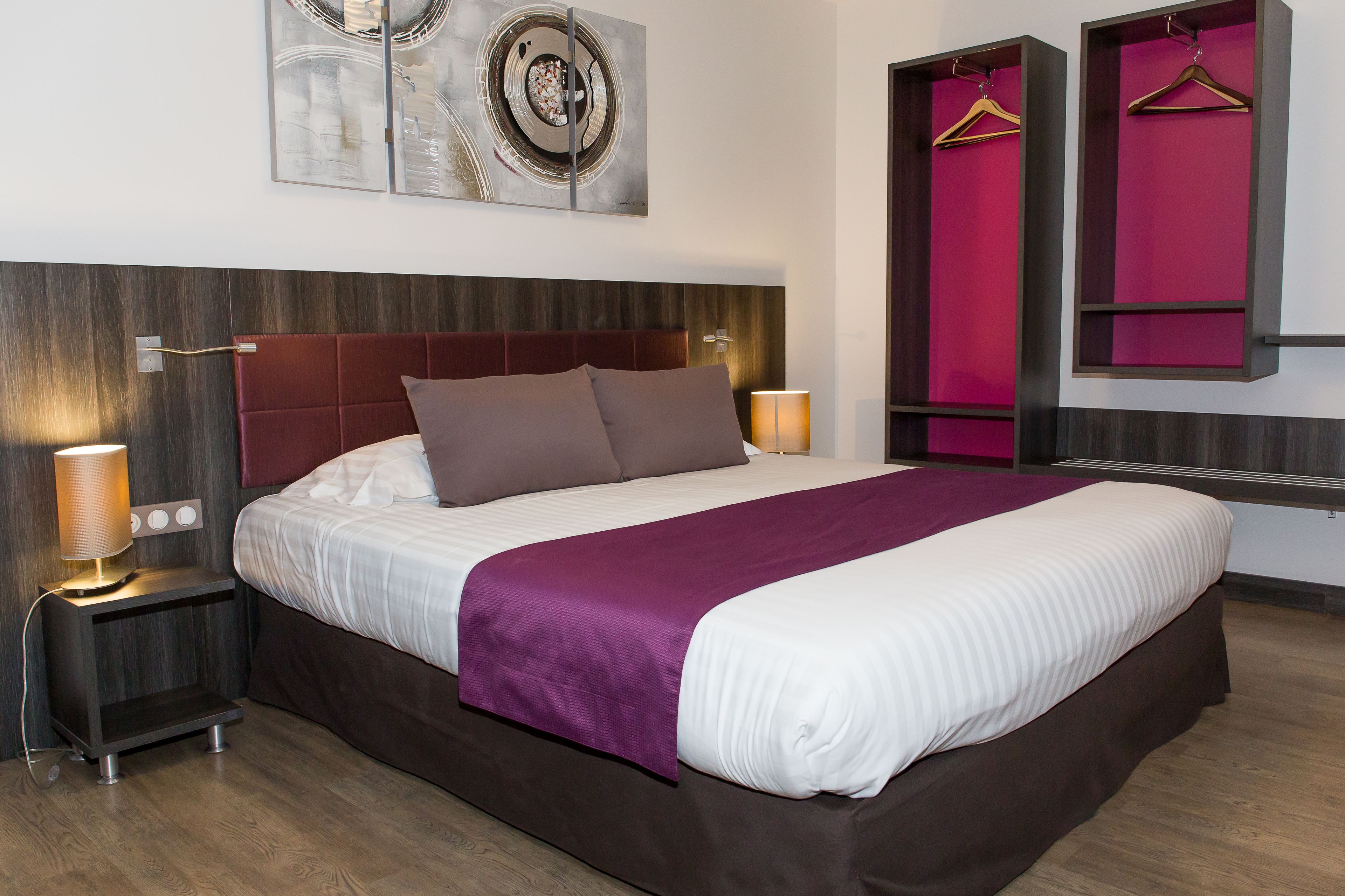 A modern hotel room with a large bed, a headboard, bedside lamps, a wall-mounted artwork, and a wardrobe with hanging space.