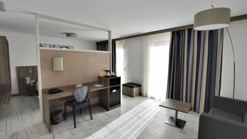 Una habitación de hotel moderna con escritorio, silla y sofá. Cuenta con una kitchenette pequeña, un cesto de basura y un ventanal grande con cortinas.