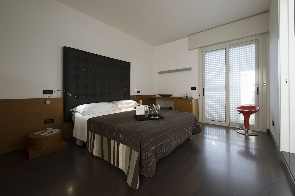 A modern hotel room with a large bed, a bedside table with a bottle and glasses, a red chair, and a sliding glass door to the outside.