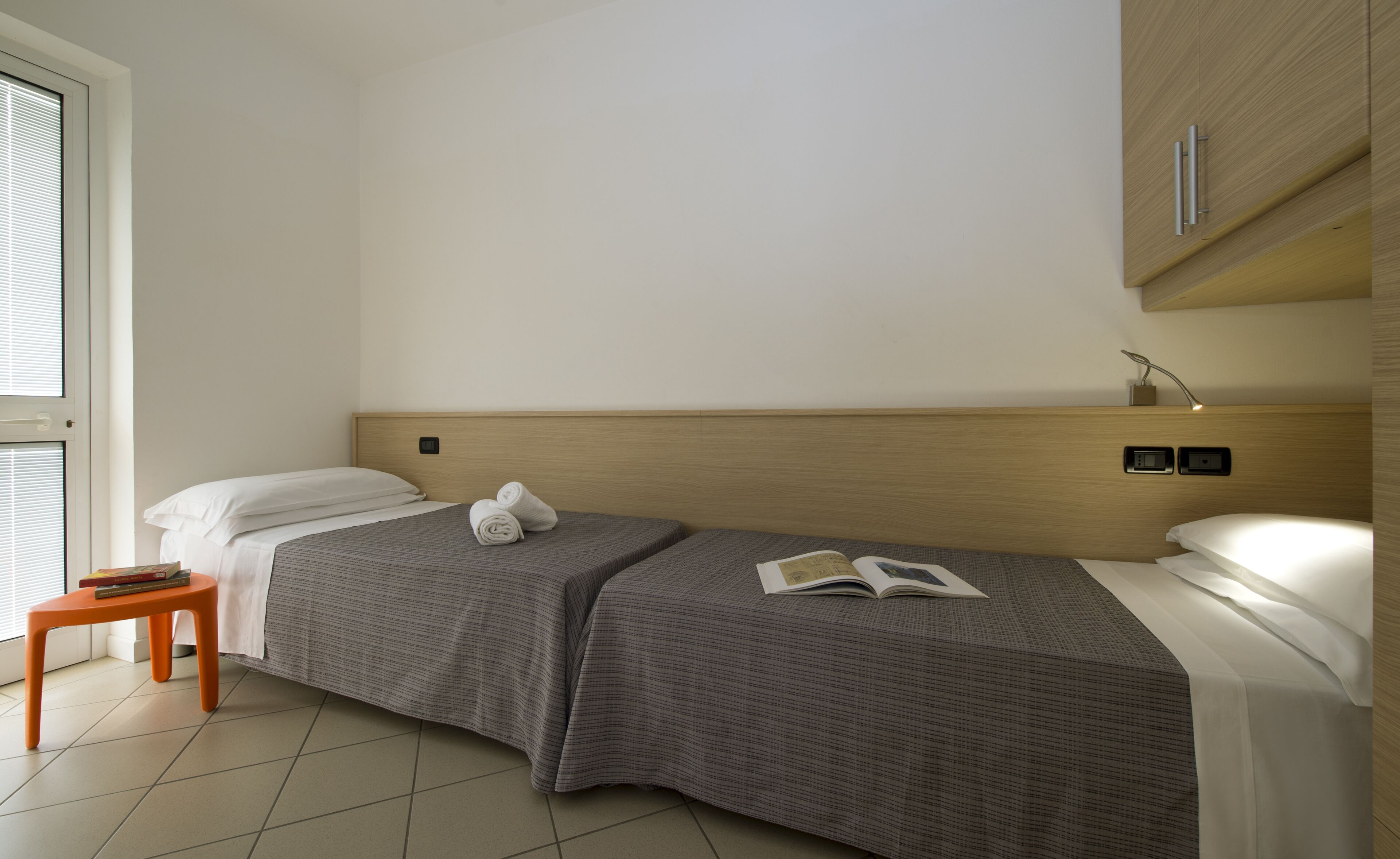 A small, neatly arranged bedroom with a single bed, a bedside table with a book, and a window with blinds.