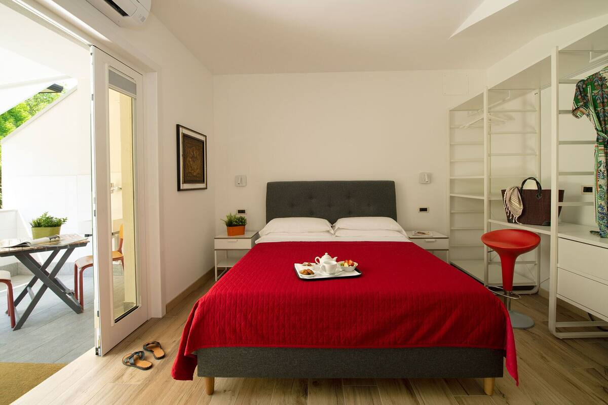A bedroom with a bed, a desk, a chair, a wardrobe, and a small table with a plant.