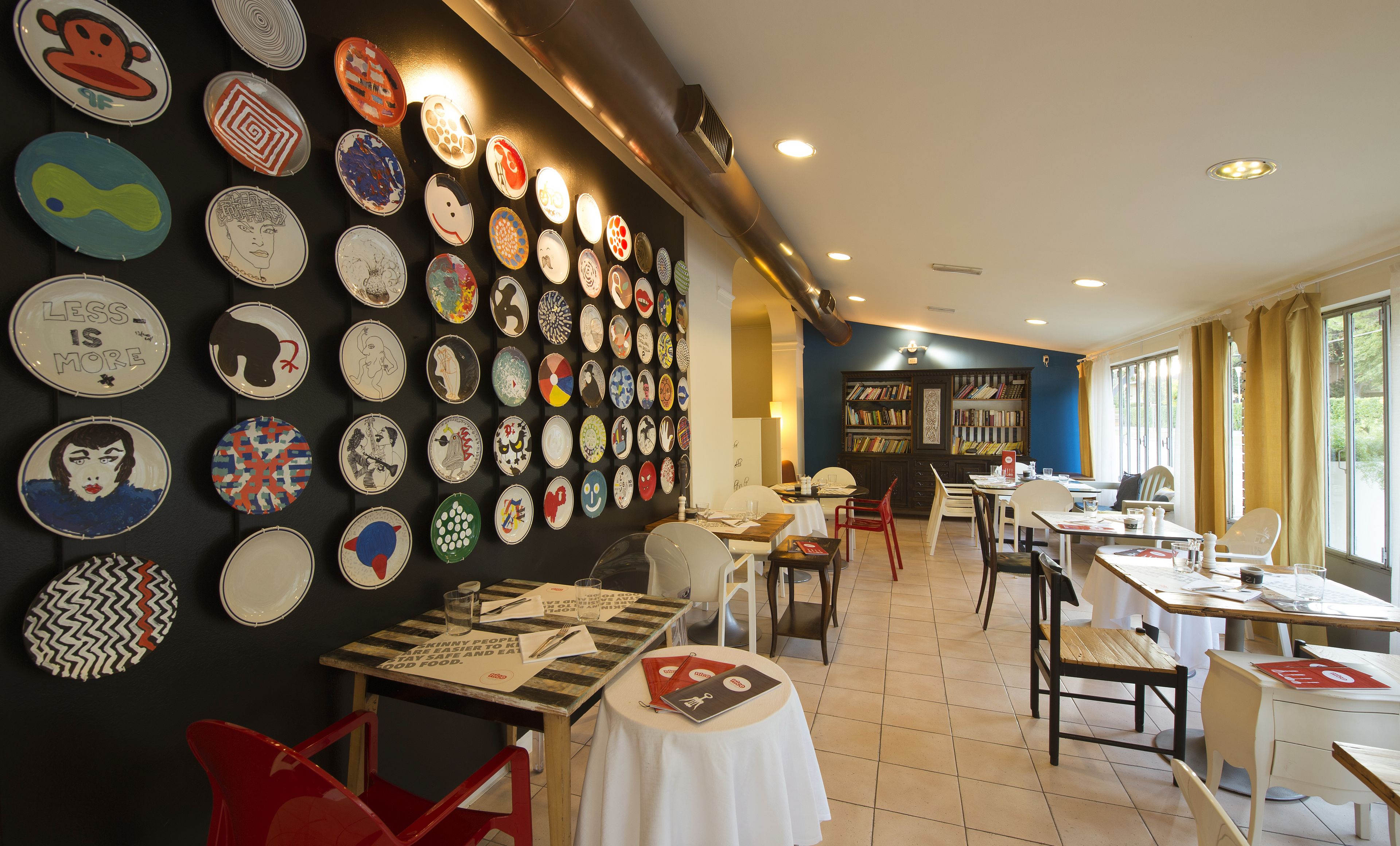 A restaurant with a wall of colorful plates, tables with menus, and a bookshelf in the background.
