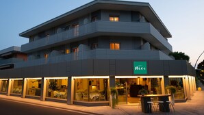 A multi-story hotel with a modern design, featuring balconies on each floor and a visible restaurant area with outdoor seating. - Nice Hotel Bibione (San Michele al Tagliamento)