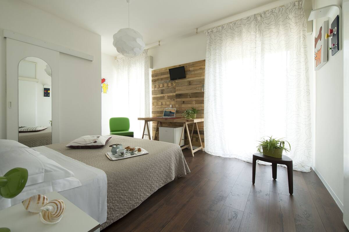A bedroom with a bed, a desk, a chair, a small table with a plant, and a mirror.