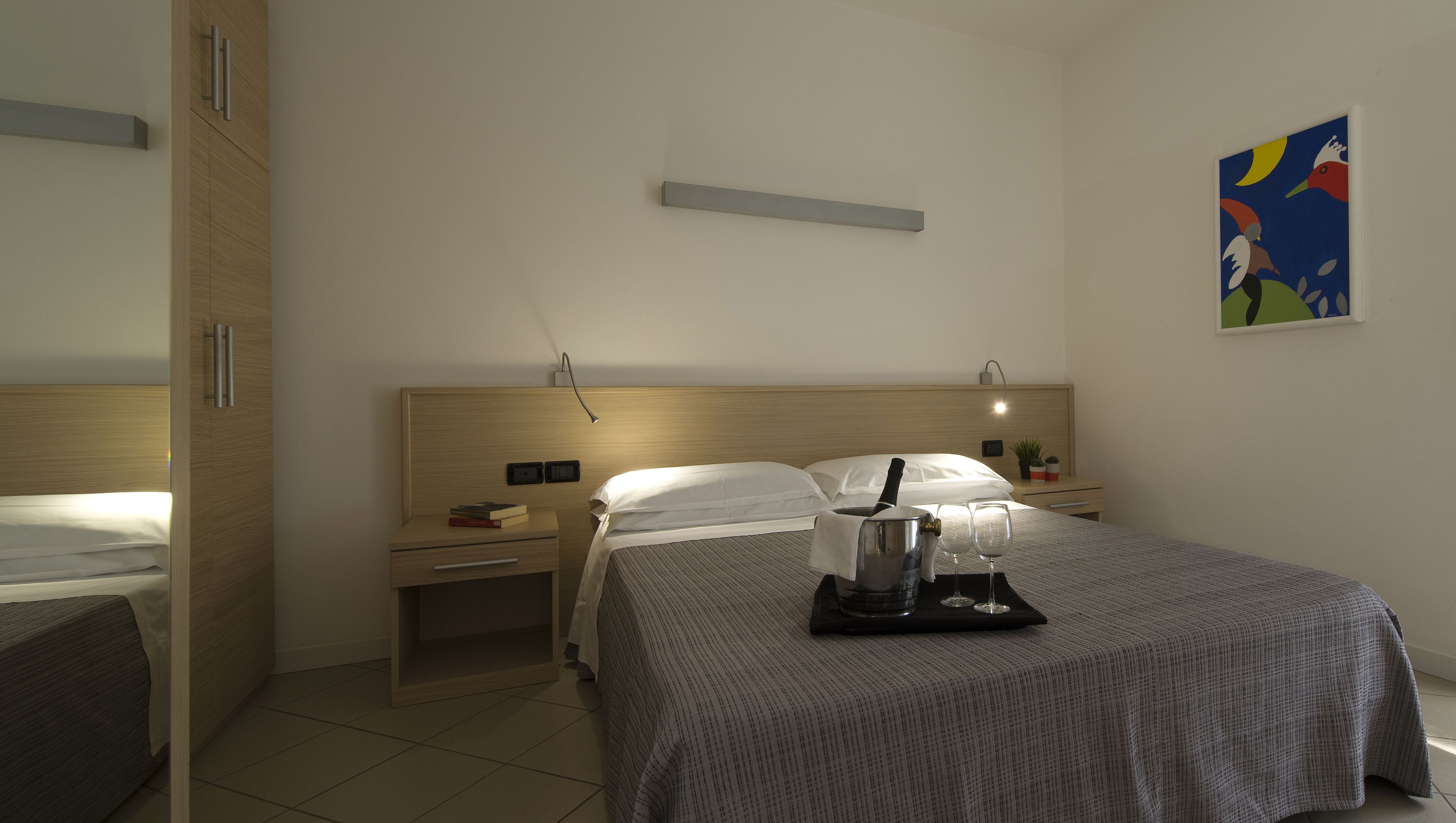 A hotel room with a bed, bedside tables, a nightstand, a lamp, a painting, and a tray with glasses and a pitcher.