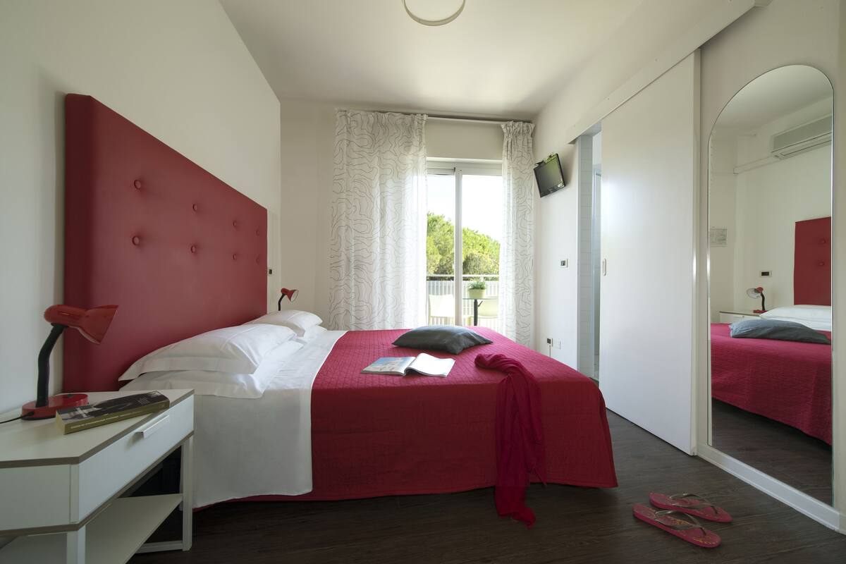 A bedroom with a bed, a desk, a mirror, and a window with curtains.