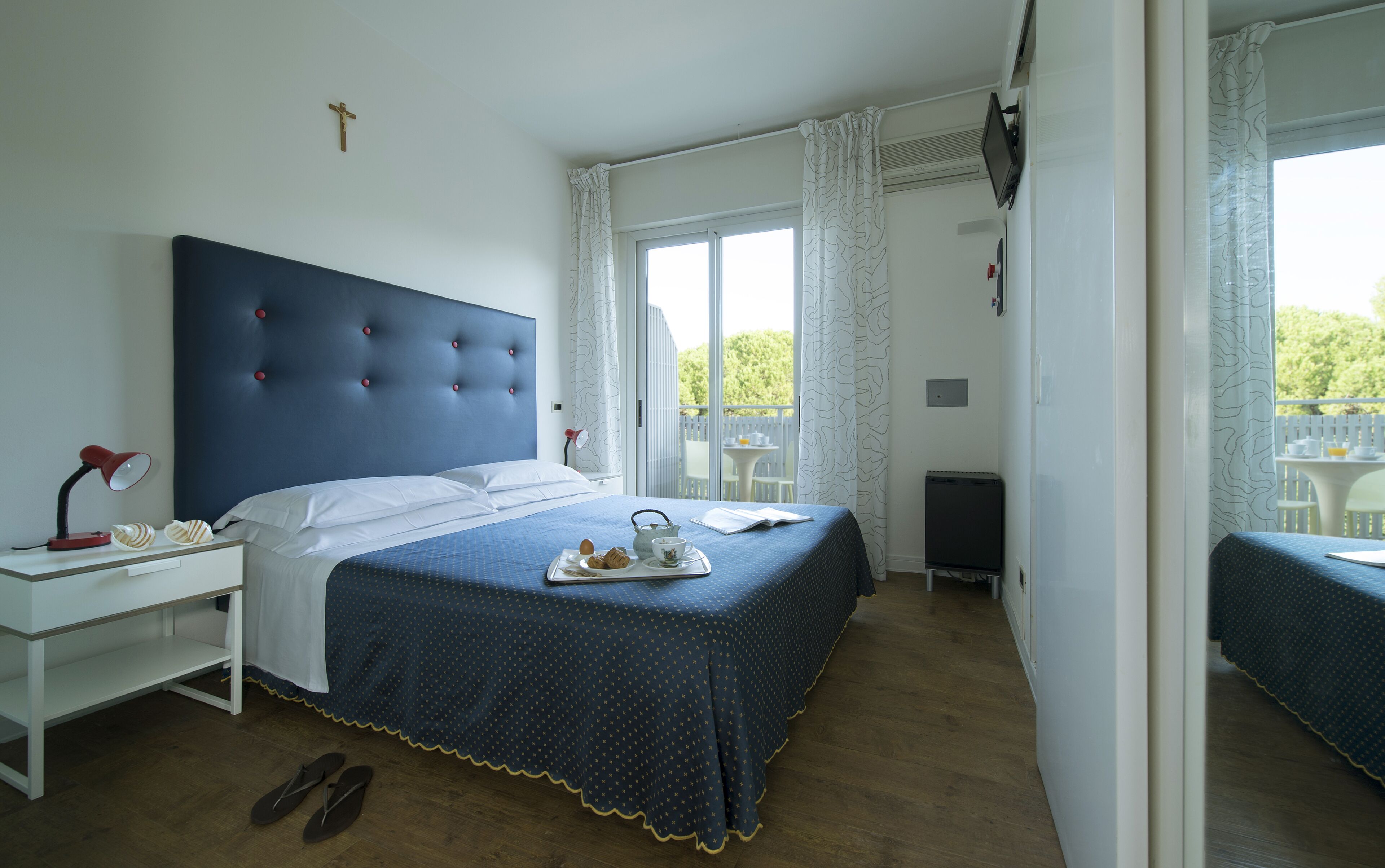Ein Schlafzimmer mit einem großen Bett, einem Nachttisch, einem Schreibtisch und einem Stuhl. Es gibt ein Fenster mit Vorhängen, einen Fernseher und einen kleinen Tisch draußen.