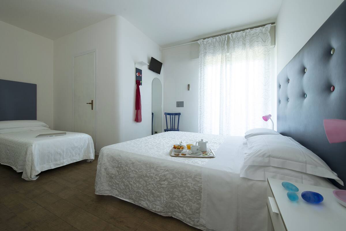 A hotel room with two single beds, a large bed with a tray of breakfast, a television, and a window with curtains.