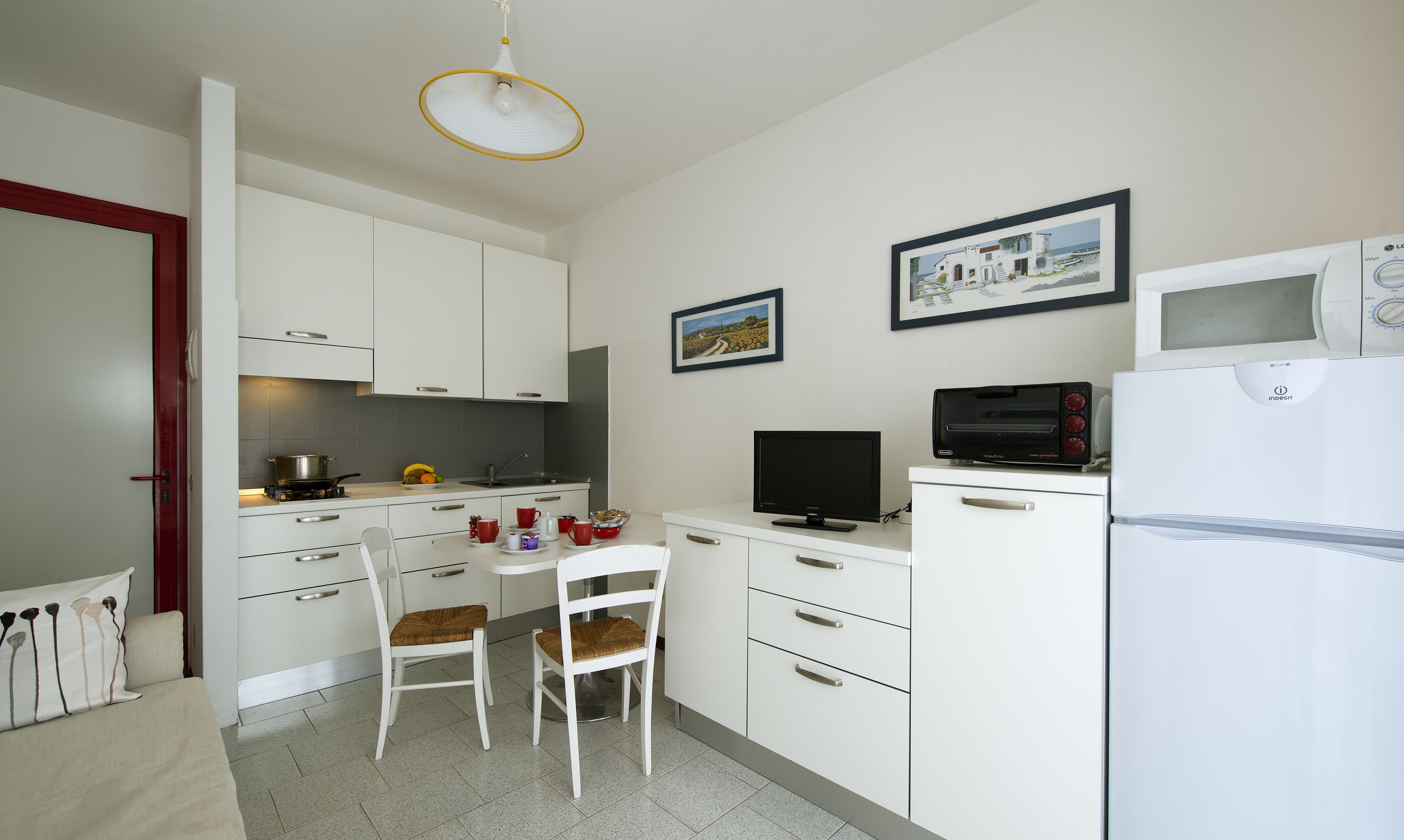 Een compacte keuken met witte kastjes, een eettafel met twee stoelen, een televisie, een magnetron en een koelkast.
