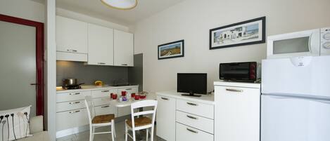 Een compacte keuken met witte kastjes, een eettafel met twee stoelen, een televisie, een magnetron en een koelkast.