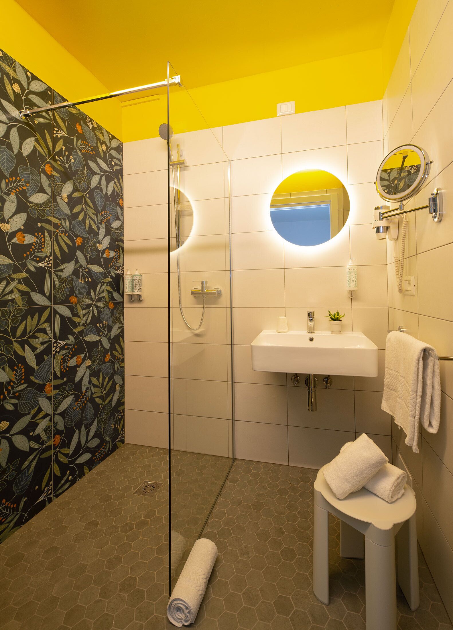 A bathroom with a glass shower enclosure, a white sink with a mirror above it, and a small stool with a towel.