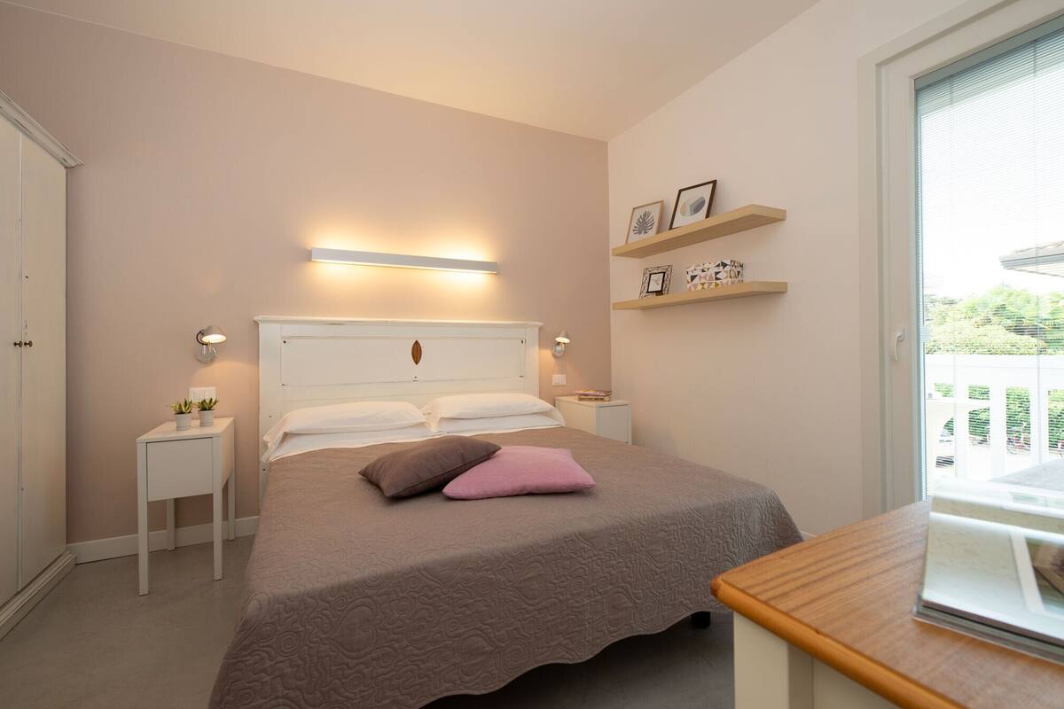 A bedroom with a bed, bedside tables, a nightstand, a window with blinds, and wall-mounted shelves.
