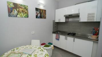 Una cocina compacta con gabinetes blancos, extractor de acero inoxidable y salpicadero de azulejos. Hay dos lámparas empotradas en la pared y un pequeño comedor con mantel estampado.