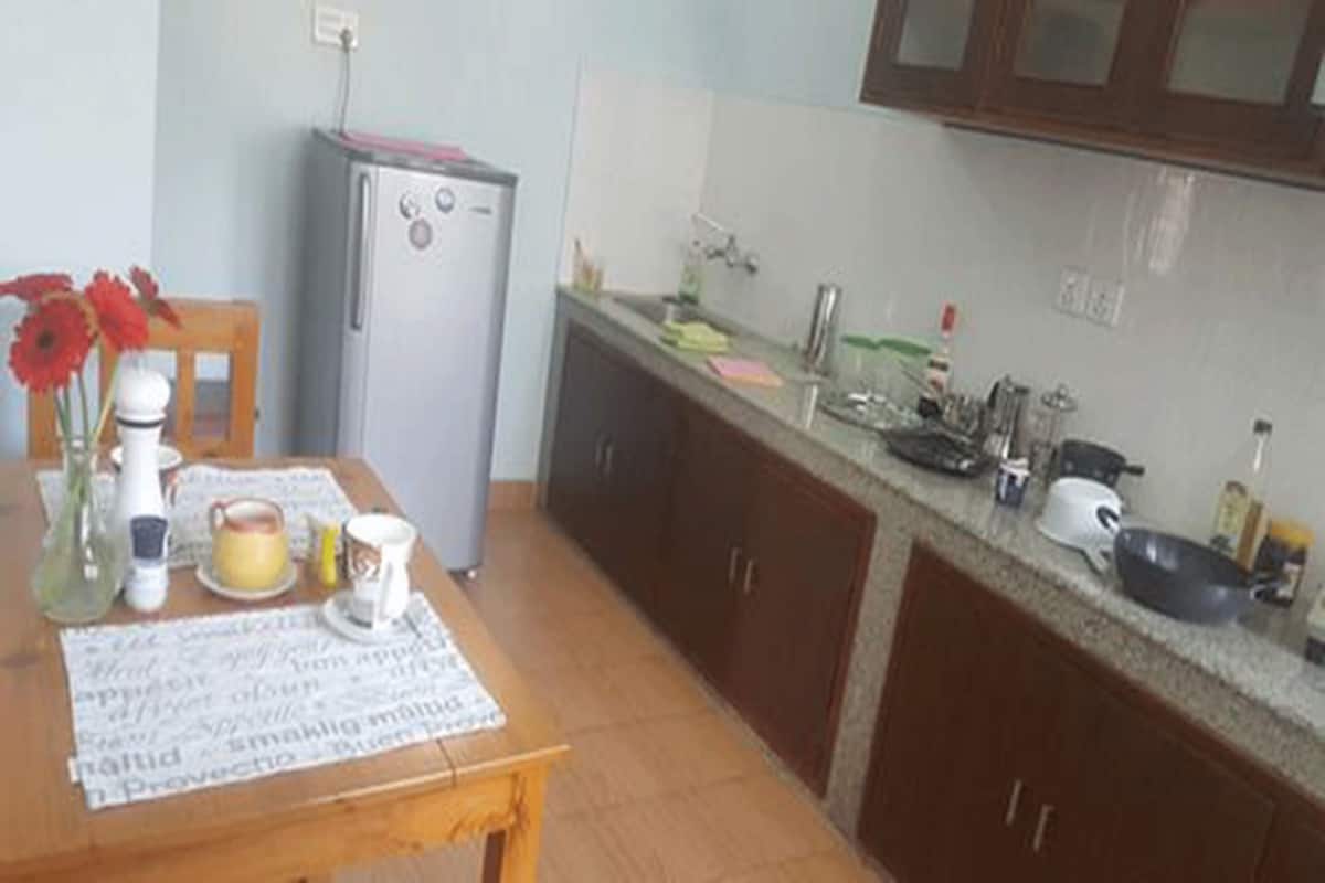 Una cocina con refrigerador, armarios de madera, una encimera con diversos utensilios y una mesa con un mantel individual.