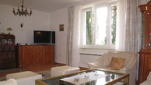Un soggiorno con tavolino in vetro, divani bianchi, televisione, armadietti in legno e un'ampia finestra con tende.