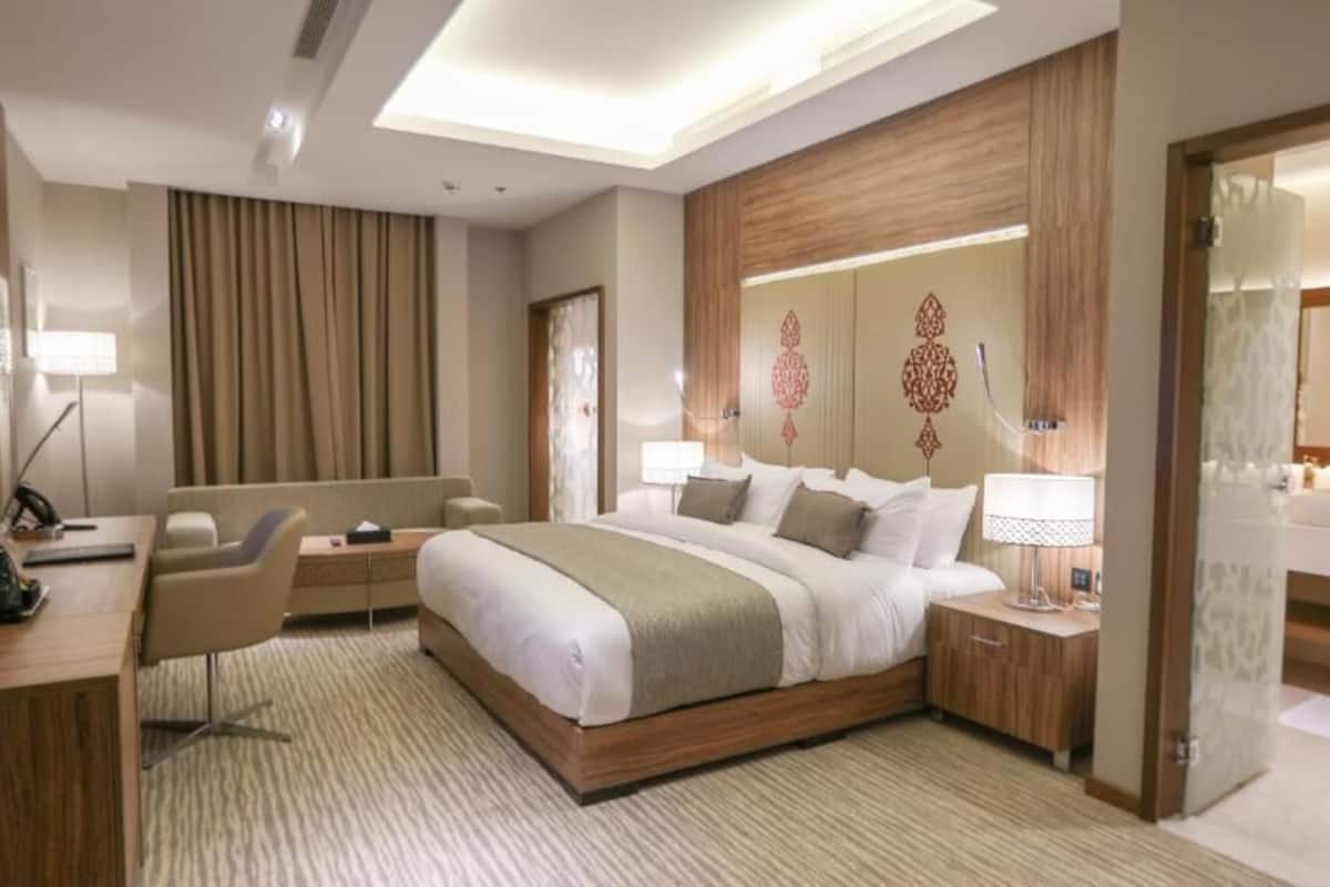A modern hotel room with a large bed, a desk with a chair, a vanity with a mirror, and a television on the wall.
