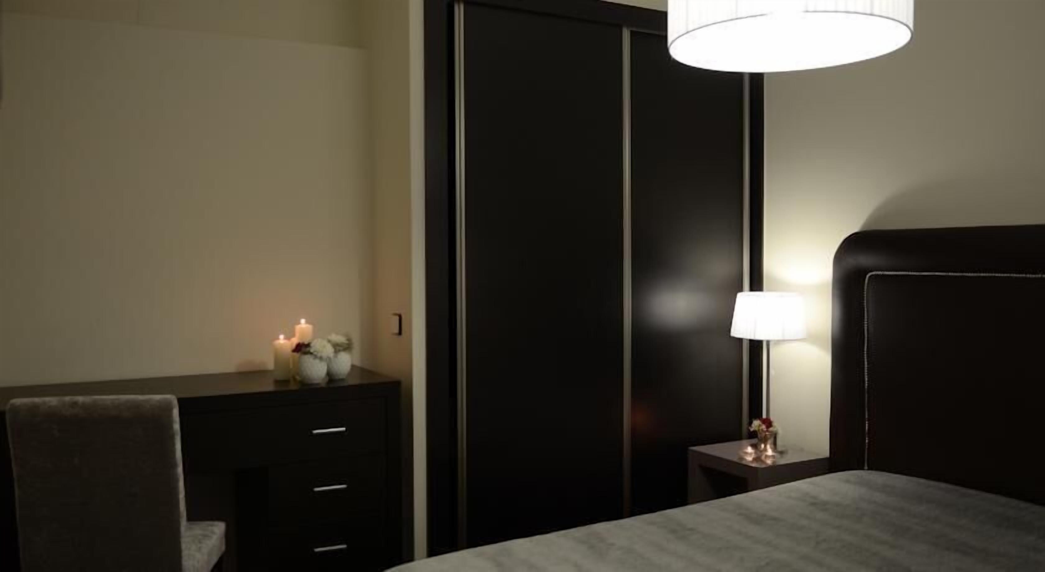 A hotel room with a bed, a bedside table with a lamp, a small table with candles, and a wardrobe.
