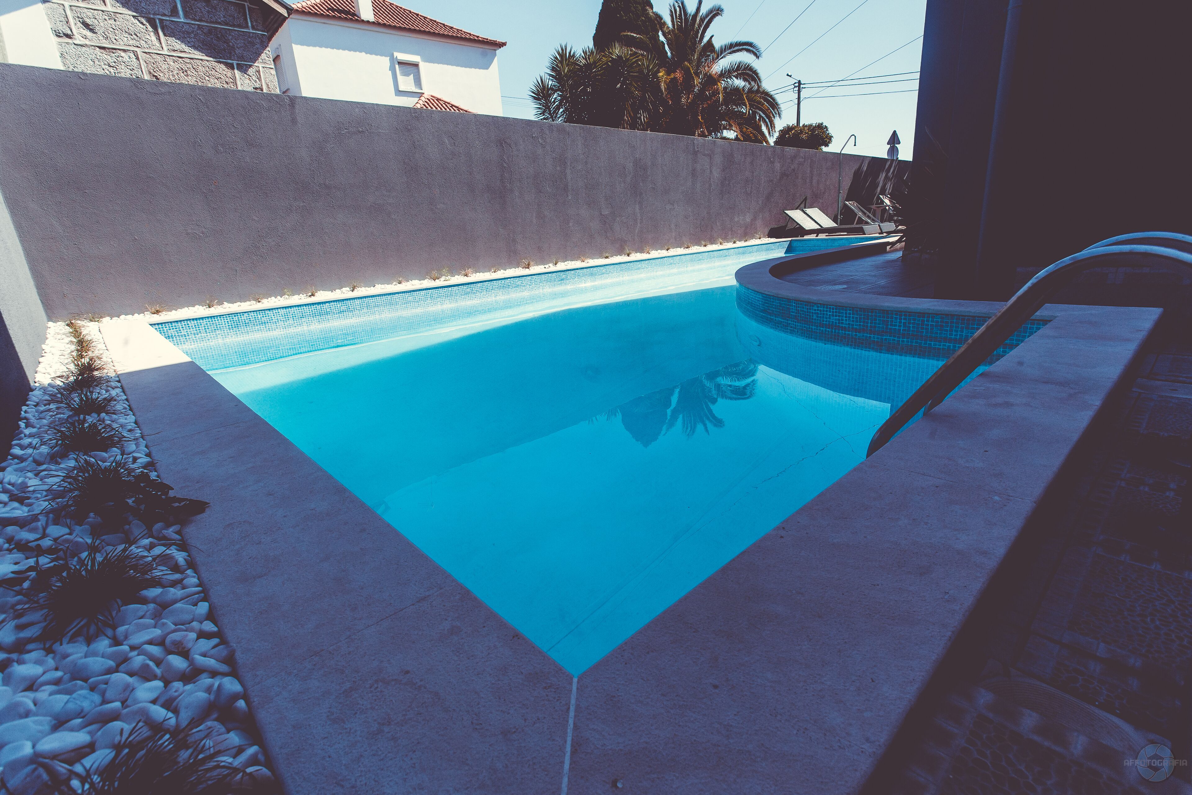 Una piscina con agua azul cristalina, rodeada por una solana de hormigón y un bajo muro de piedra.