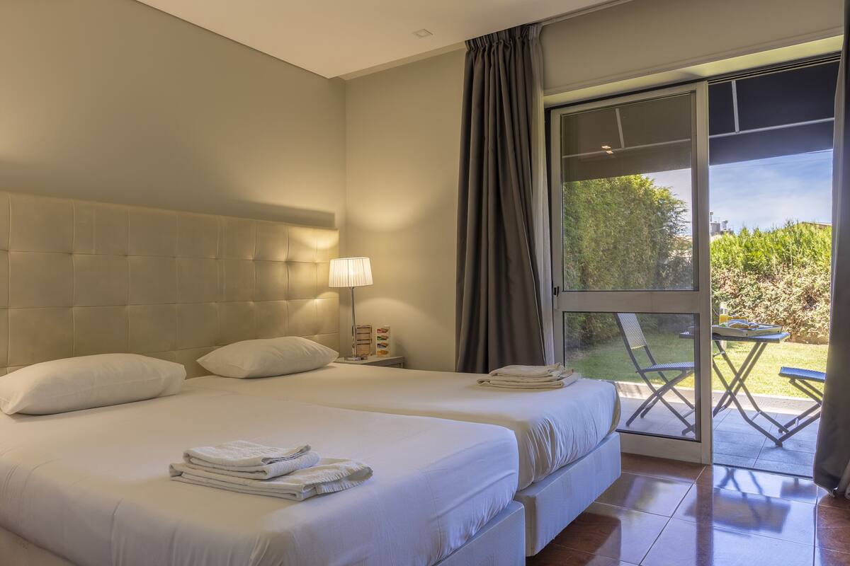 A hotel room with two beds, a nightstand with a lamp, and a view of the outdoors through a sliding glass door.