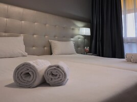 Habitación de hotel con una cama grande, dos almohadas, una mesita de noche con una lámpara y una toalla enrollada sobre la cama.