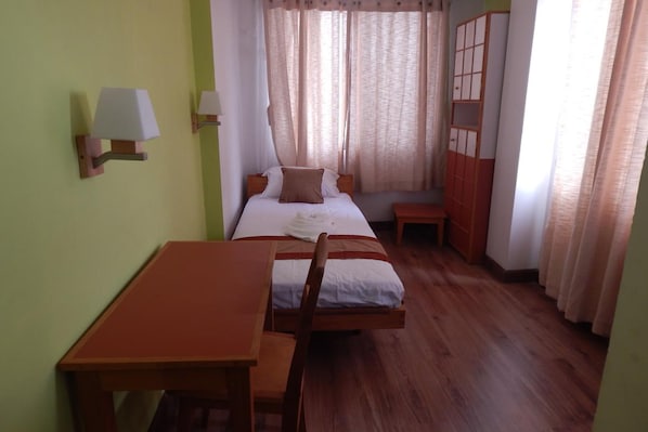 A small, neatly arranged room with a bed, a wooden table, and a chair. There are two wall-mounted lamps and a window with curtains.