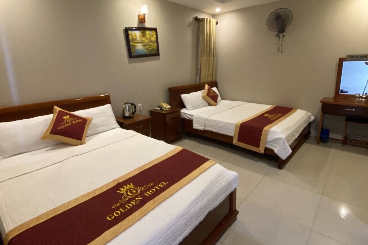 Une chambre d’hôtel avec deux lits, chacun recouvert d’une courtepointe rouge et or, une table de chevet, une lampe, un tableau au mur et un ventilateur de plafond.