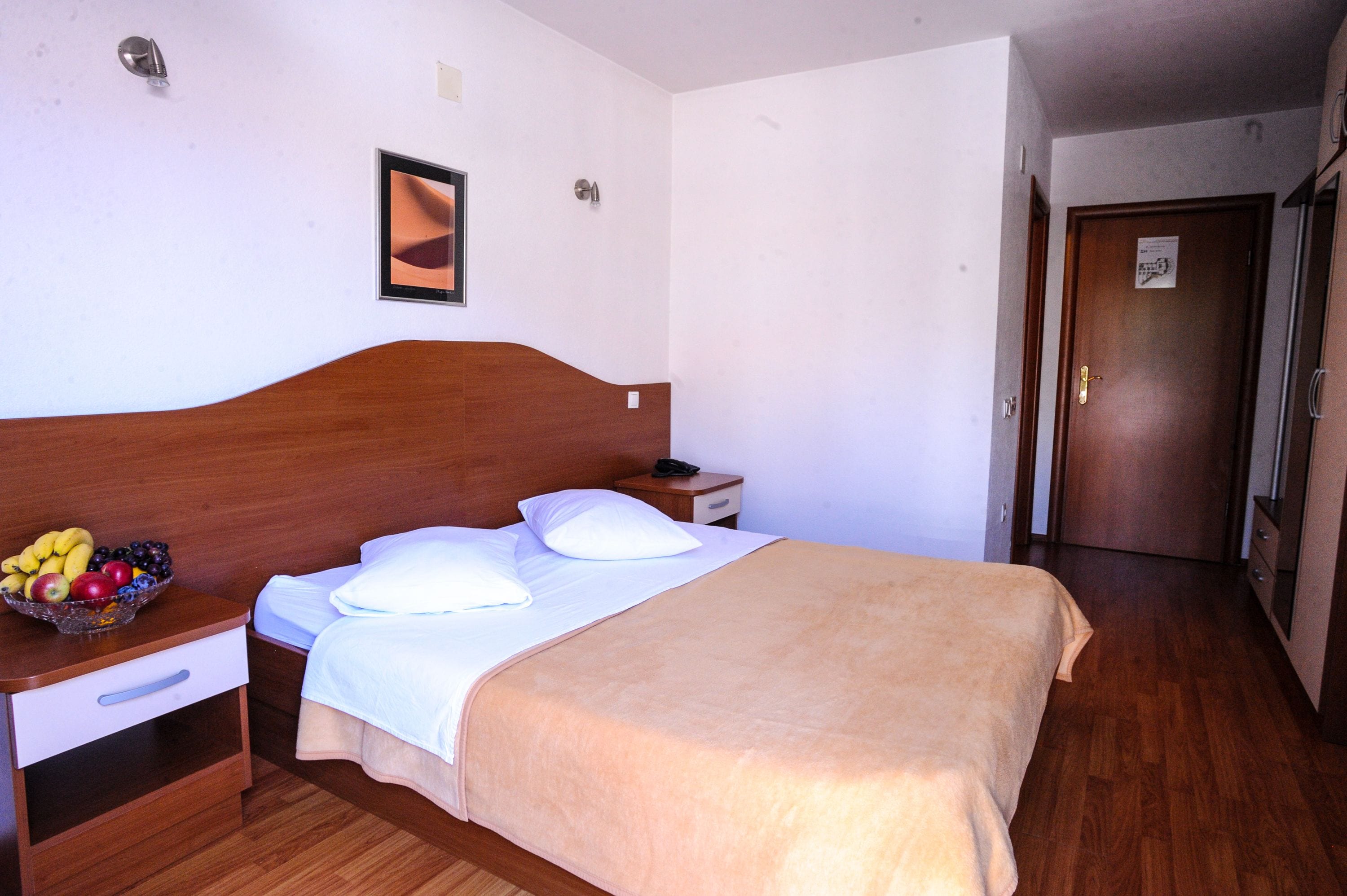 Una camera d'albergo con testiera in legno, un letto con lenzuola bianche, una coperta color beige, un comodino con una ciotola di frutta e un tavolino con il telefono.