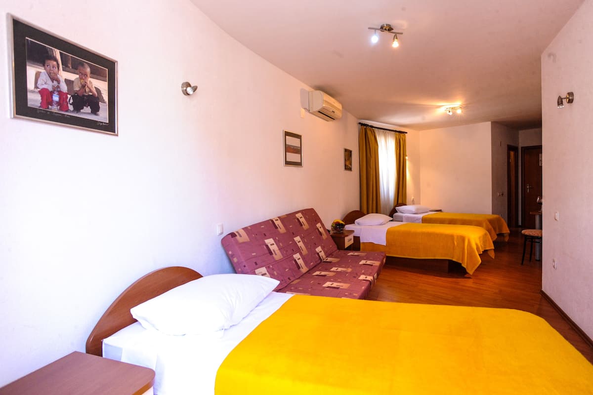 Quarto de hotel com sofá-cama, duas camas de solteiro, uma cadeira de madeira, uma mesinha e uma fotografia emoldurada na parede.