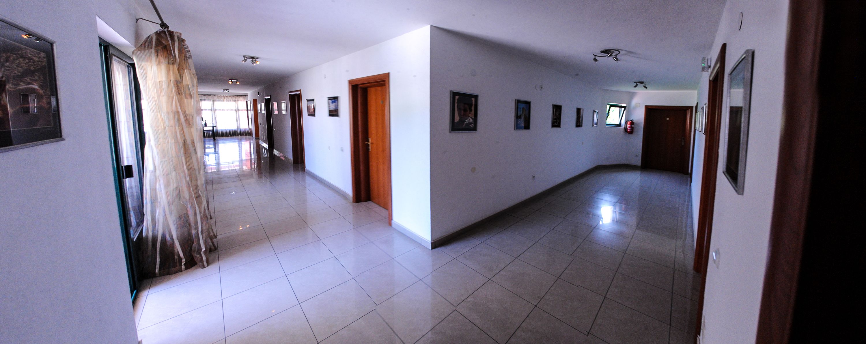 A long, narrow hallway with tiled floors, wooden doors, and framed pictures on the walls.