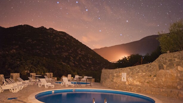 Une piscine entourée de chaises longues et d’un mur de pierre, le tout surplombé par des montagnes et baigné par un ciel étoilé.