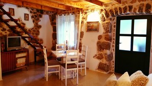 Un salon chaleureux avec un mur en pierre, un plafond en bois, un coin repas avec une table et des chaises, un téléviseur posé sur un meuble en bois et un escalier.