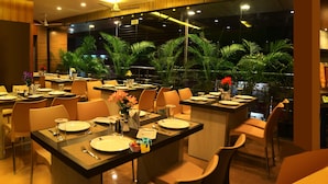Un restaurante moderno con varias áreas de comedor, que incluye un bar y una zona de estar con vistas a plantas tropicales.