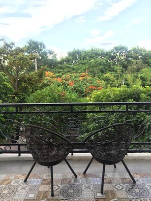 A balcony with two ornate metal chairs, a tiled floor with a geometric pattern, and a lush garden with various plants and flowers in the background. - Cochi Villa Hoi An (Da Nang)