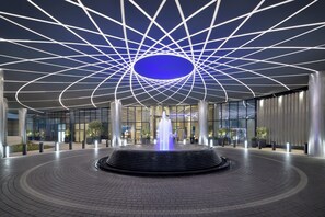 A modern building entrance with a geometric ceiling design, a central fountain, and a paved walkway.