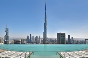 A view of a modern cityscape with a prominent skyscraper, a clear blue sky, and a swimming pool with lounge chairs.