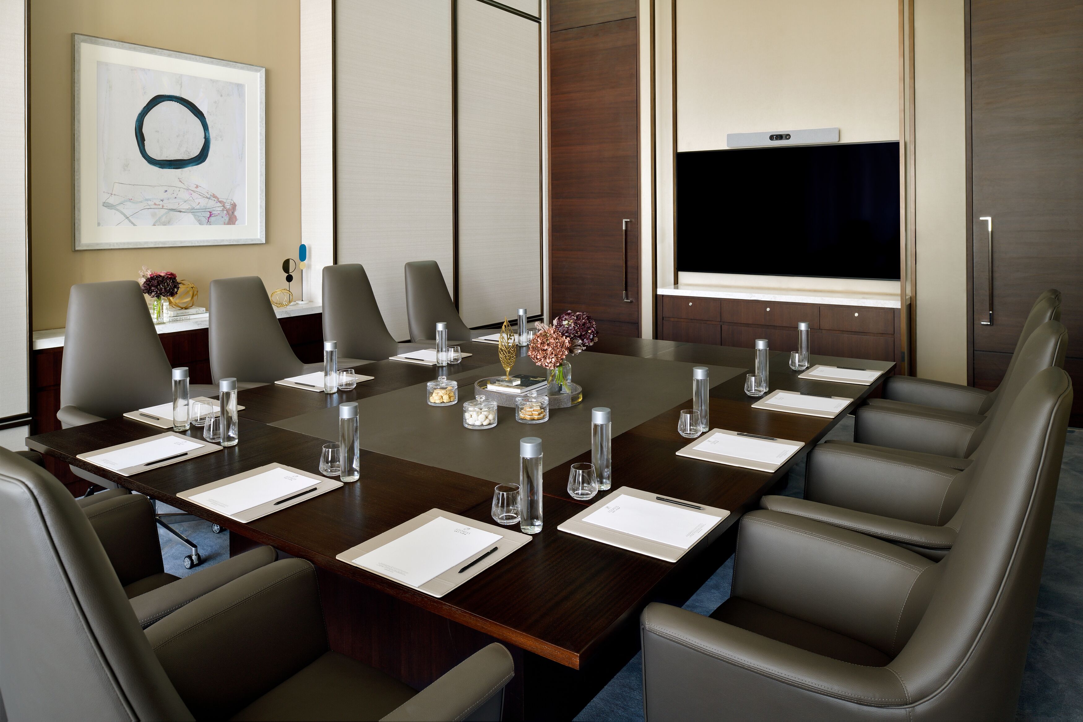 A conference room with a long table, multiple chairs, a flat-screen TV, and a framed abstract artwork on the wall.