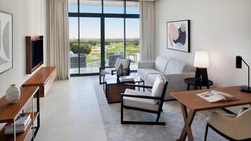 Et moderne hotelværelse med et stort vindue der giver udsigt over en golfbane, en sofa, et sofabord, et fjernsyn og et skrivebord med en lampe.