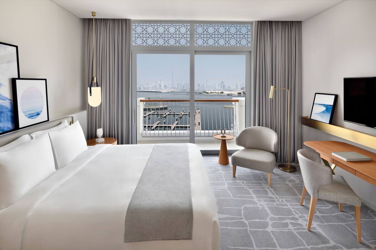 A hotel room with a large bed, a desk, a chair, a TV, and a view of the city skyline through a window.