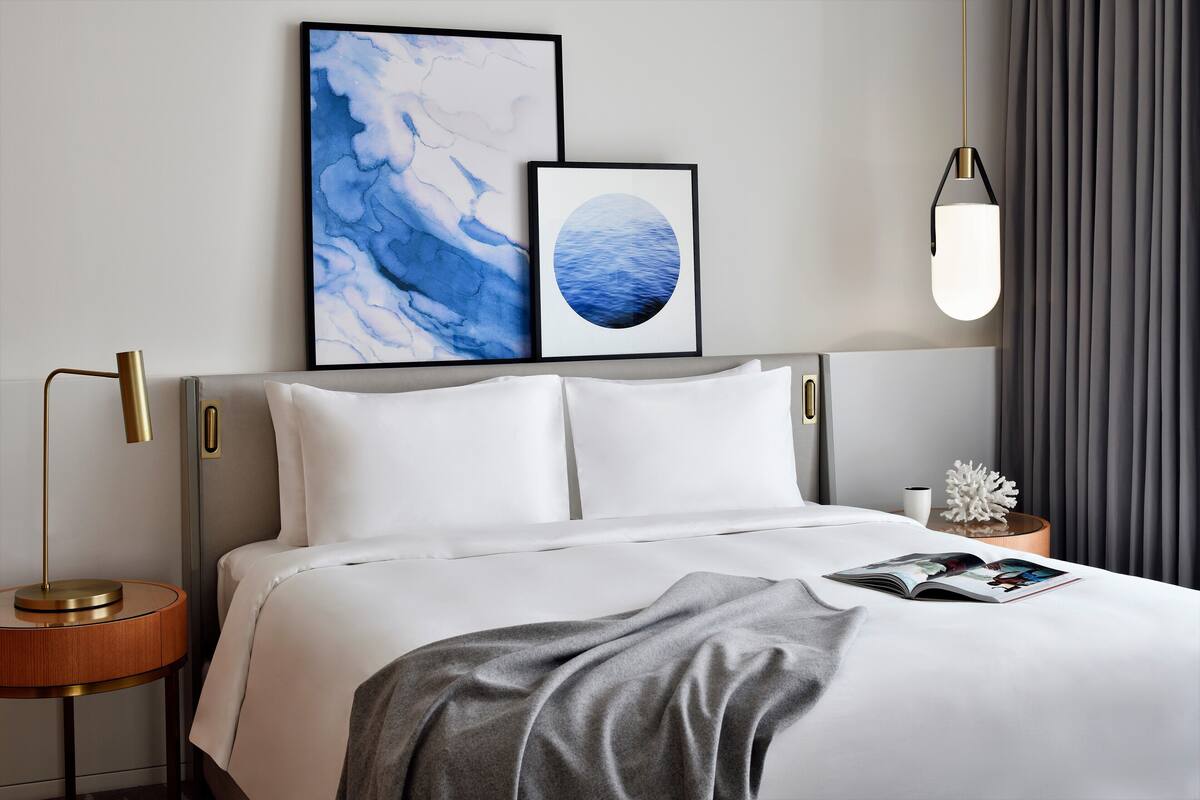 A neatly made bed with white linens, a grey throw blanket, and a magazine on the bed. There is a framed artwork above the bed, a bedside lamp, and a decorative coral piece on a nightstand.