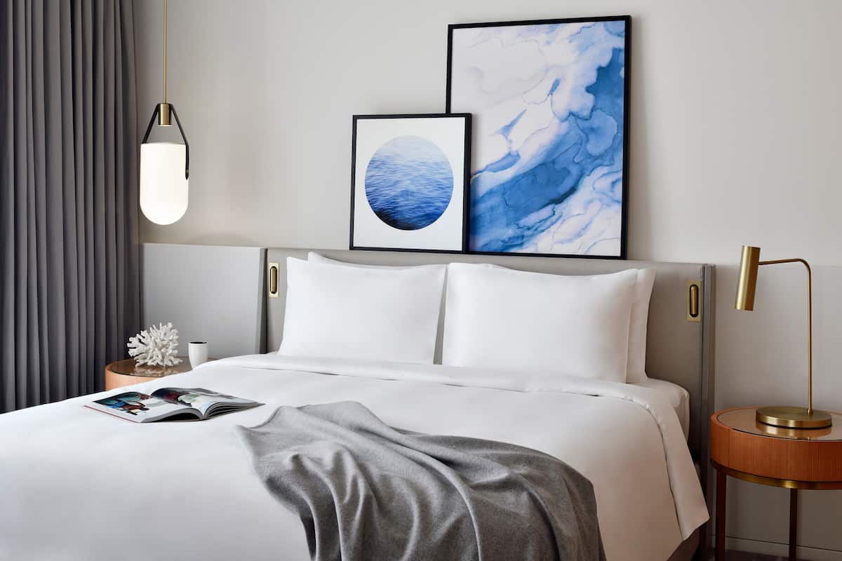 A modern hotel room with a neatly made bed, a bedside table with a lamp, and two framed artworks on the wall.