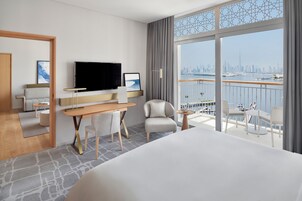 A modern hotel room with a large bed, a desk with a flat-screen TV, a comfortable armchair, and a balcony with a city view.