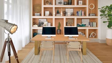 A room with two monitors on a wooden table, a tripod with a lamp, and a bookshelf filled with books and decorative items.