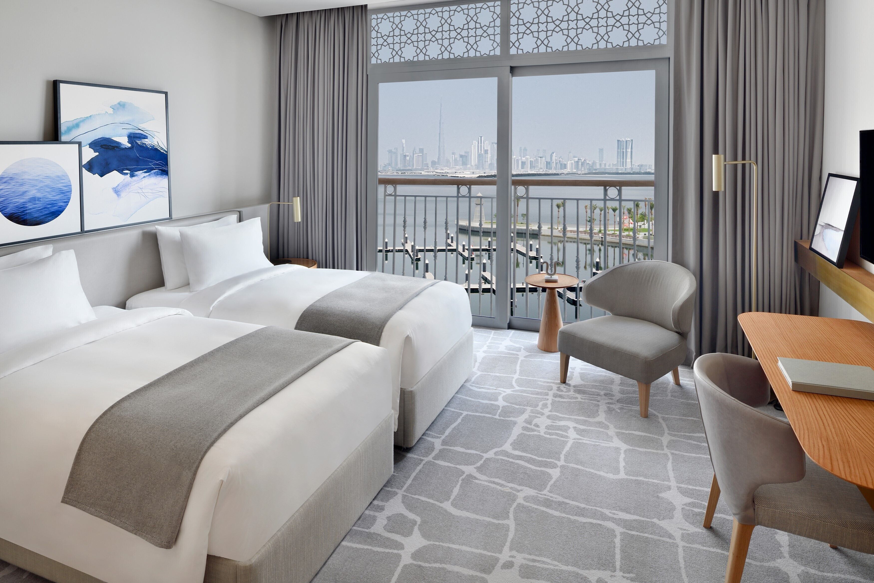 A hotel room with two beds, a desk, a chair, and a balcony with a city view.